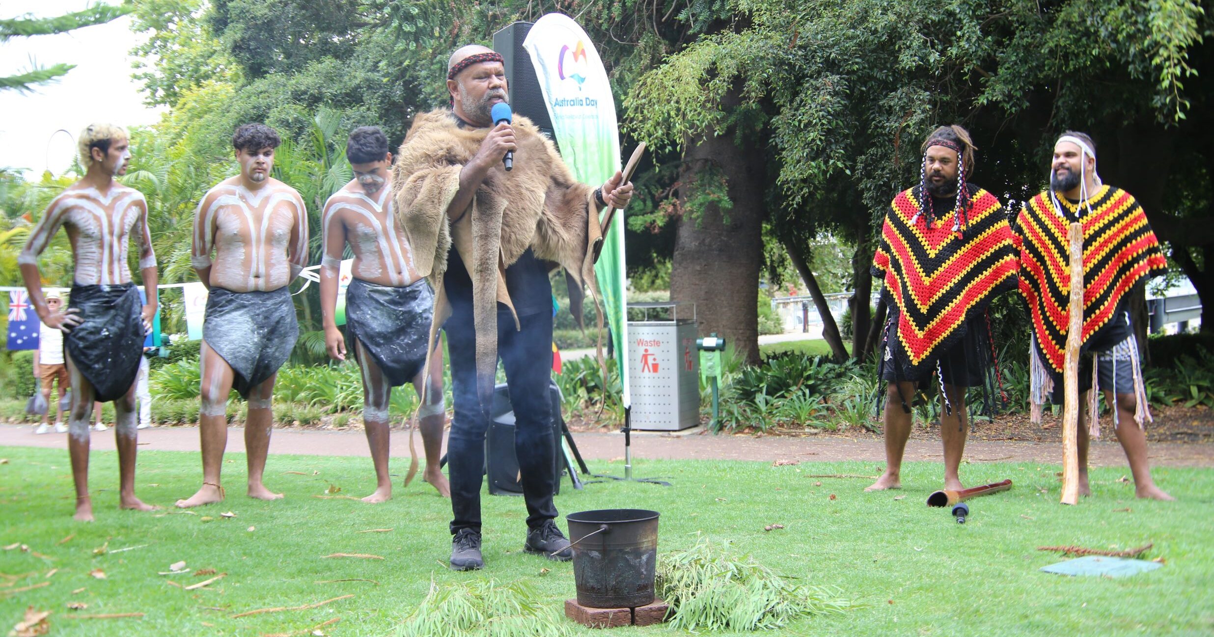 Aboriginal man speaking into microphone with dancers behind wearing traditional clothing