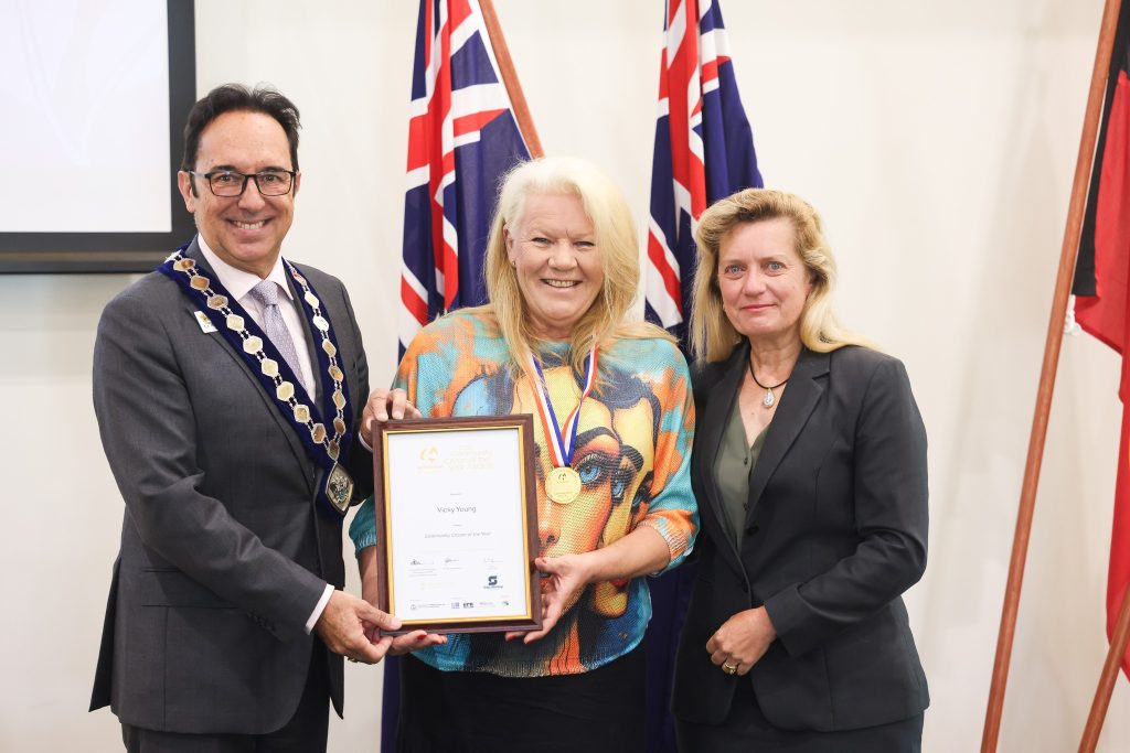 Three people smiling to camera in front of Australian flags, middle person holding certificate and wearing a medallion
