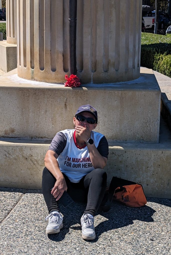 Patchara Weggers sitting on the ground in front of column and a bunch of poppies, wearing a t-shirt that says I'm Marching For Our Heroes